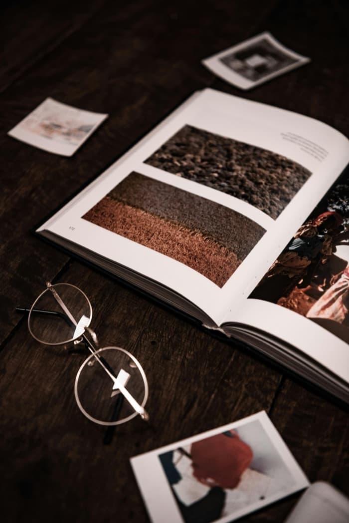 An open photobook on a dark wooden table beside round glasses and loose prints